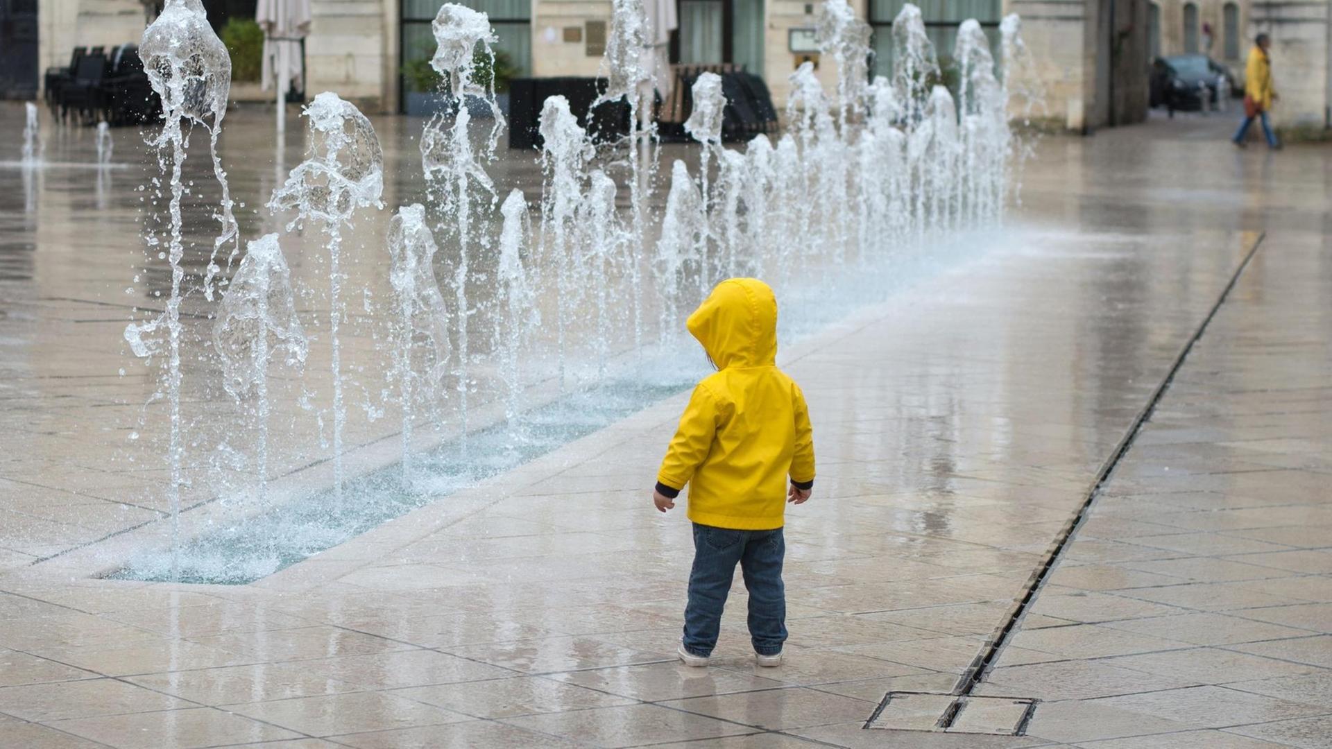 Kleinkind in gelber Regenjacke steht vor einem Springbrunnen