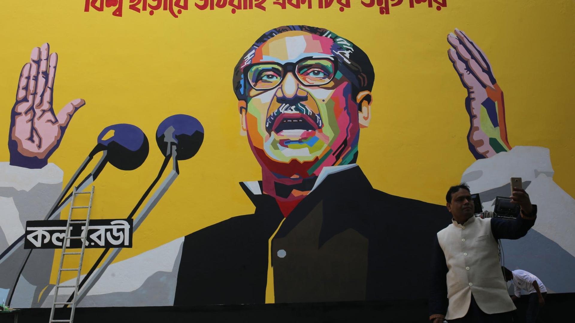 Ein Farbfoto zeigt einen Mann, der sich mit seinem Handy vor einer riesigen Plakatwand ablichtet, auf der Mujibur Rahman In staatstragender Geste abgebildet ist abgebildet ist Ein Farbfoto zeigt einen Mann, der sich mit seinem Handy vor einer riesigen Plakatwand ablichtet, auf der Mujibur Rahman In staatstragender Geste abgebildet ist abgebildet ist