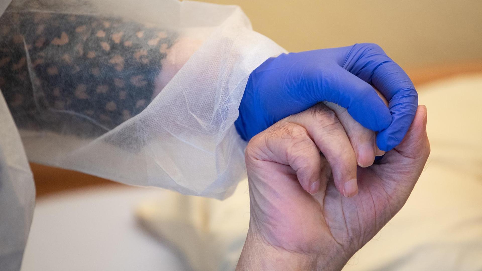18.11.2020, Baden-Württemberg, Tübingen: Eine Altenpflegerin in Schutzausrüstunghält die Hand eines Bewohners. Foto: Sebastian Gollnow/dpa | Verwendung weltweit 18.11.2020, Baden-Württemberg, Tübingen: Eine Altenpflegerin in Schutzausrüstunghält die Hand eines Bewohners. Foto: Sebastian Gollnow/dpa | Verwendung weltweit