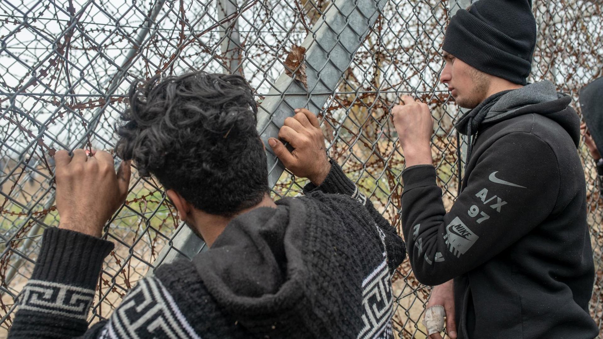 Zwei junge Männer stehen an einem Stacheldraht-Zaun und blicken hindurch.