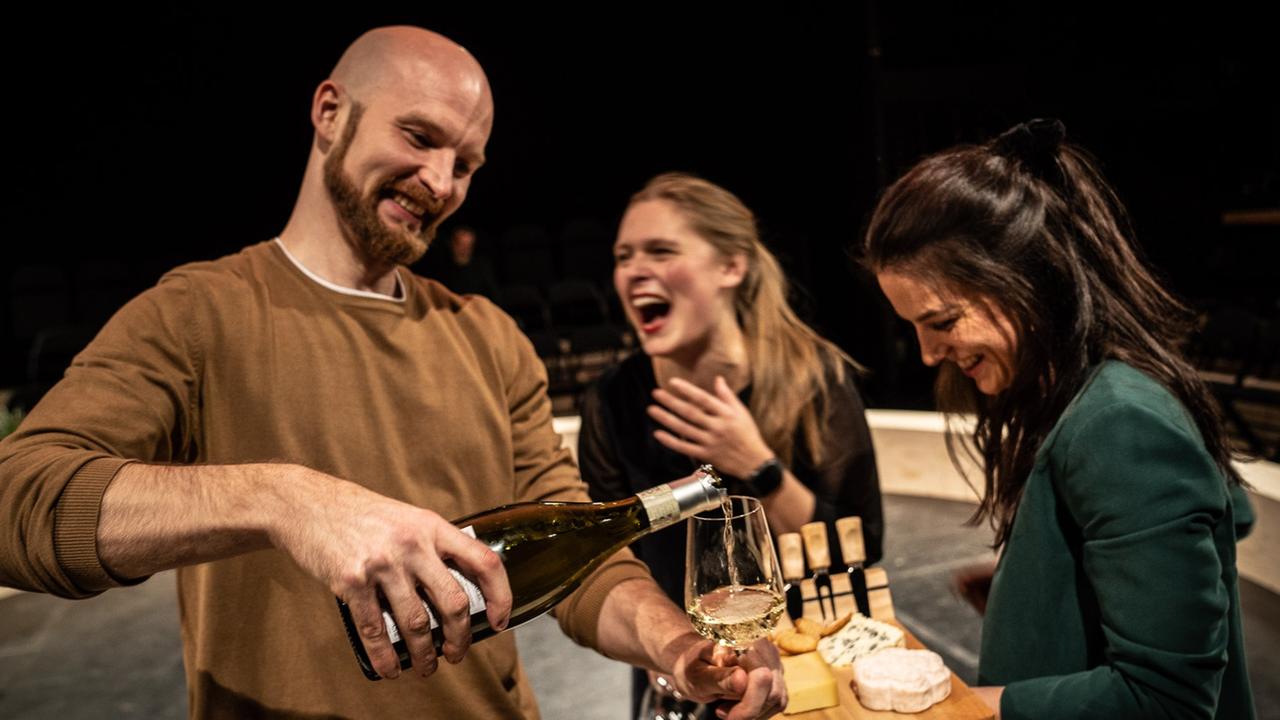 Eine Szene aus dem Theaterstück "White Male Privilege". Zu sehen sind zwei Frauen und ein Mann, alle drei stehen. Die Frau, die ganz rechts steht, hält ein Tablett mit Käse. Der Mann - zur Linken - schenkt gerade aus einer Flasche Weißwein in ein Weinglas ein. Die Frau, die in der Mitte steht, lacht herzlich. Eine Szene aus dem Theaterstück "White Male Privilege". Zu sehen sind zwei Frauen und ein Mann, alle drei stehen. Die Frau, die ganz rechts steht, hält ein Tablett mit Käse. Der Mann - zur Linken - schenkt gerade aus einer Flasche Weißwein in ein Weinglas ein. Die Frau, die in der Mitte steht, lacht herzlich.