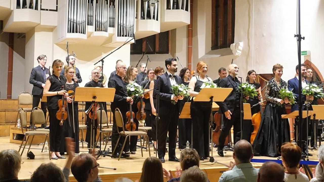 Orchester, Sänger und dirigent nehmen Beifall des Publikums stehend auf einer Bühne entgegen. Orchester, Sänger und dirigent nehmen Beifall des Publikums stehend auf einer Bühne entgegen.