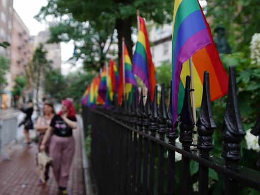 An einem Stahlzaun sind Regenbogenflaggen befestigt. In der Unschärfe laufen Frauen vorbei. Eine trägt eine pinke Frisur.