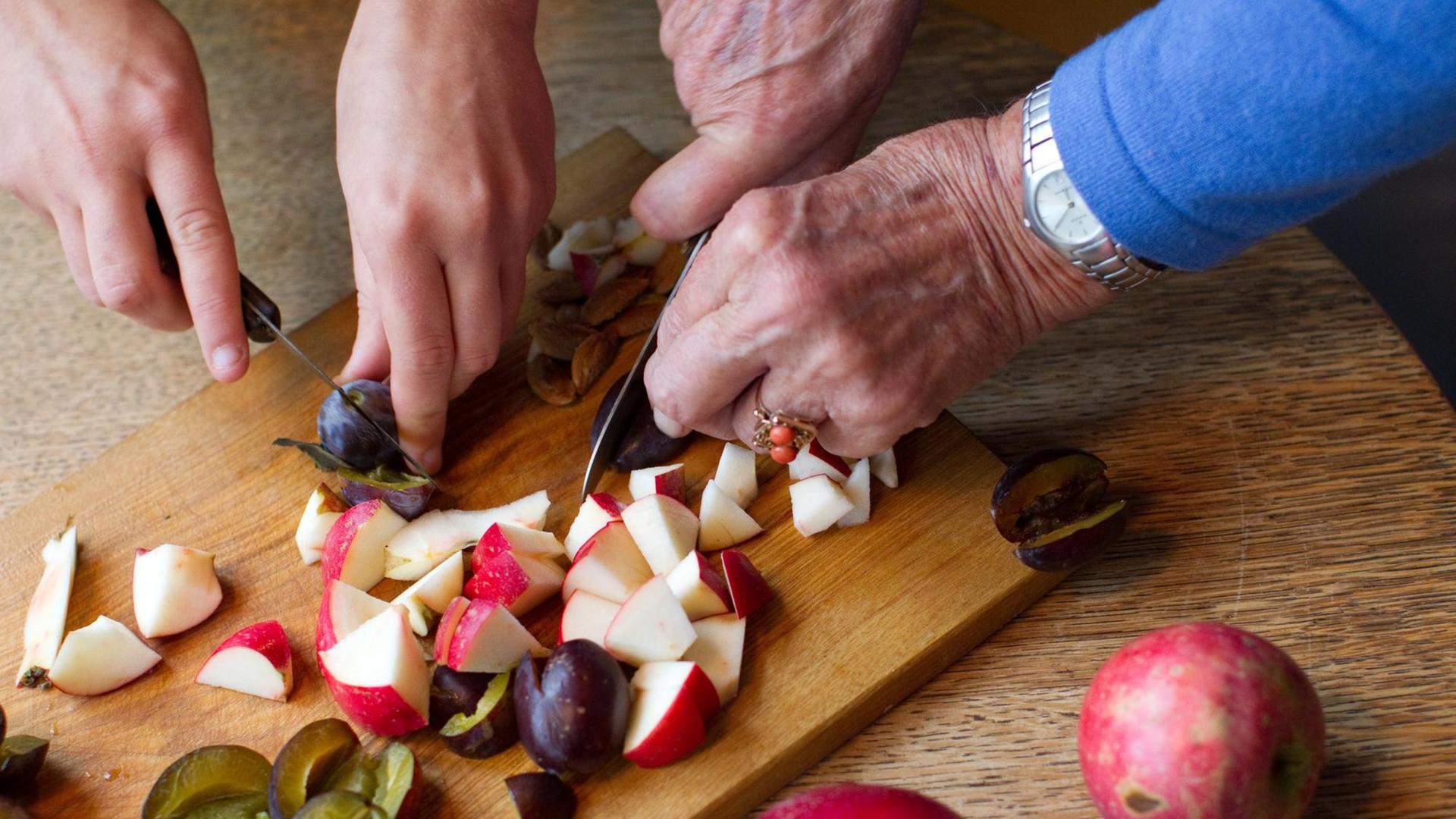 Eine Seniorin und eine Studentin schneiden gemeinsam Obst in der Küche.