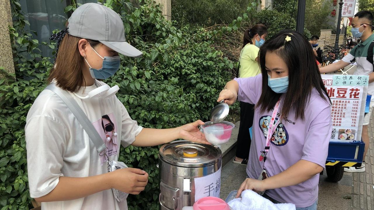 Eine junge Frau hält an einem Straßenstand ein Gefäß aus Plastik, in das eine andere Essen füllt. Eine junge Frau hält an einem Straßenstand ein Gefäß aus Plastik, in das eine andere Essen füllt.