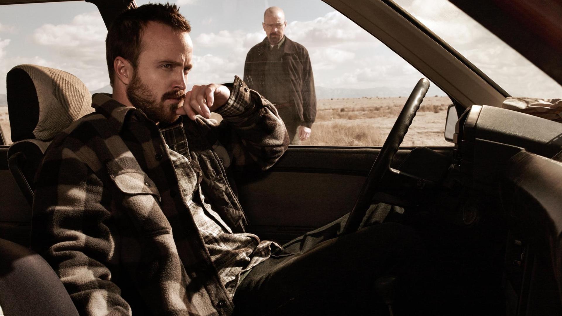 Szenenbild aus "Breaking Bad": Aaron Paul als Jessie Pinkman sitzt im Auto, im Hintergrund sieht man Bryan Cranston als Walter "Walt" Hartwell White stehen. Szenenbild aus "Breaking Bad": Aaron Paul als Jessie Pinkman sitzt im Auto, im Hintergrund sieht man Bryan Cranston als Walter "Walt" Hartwell White stehen.