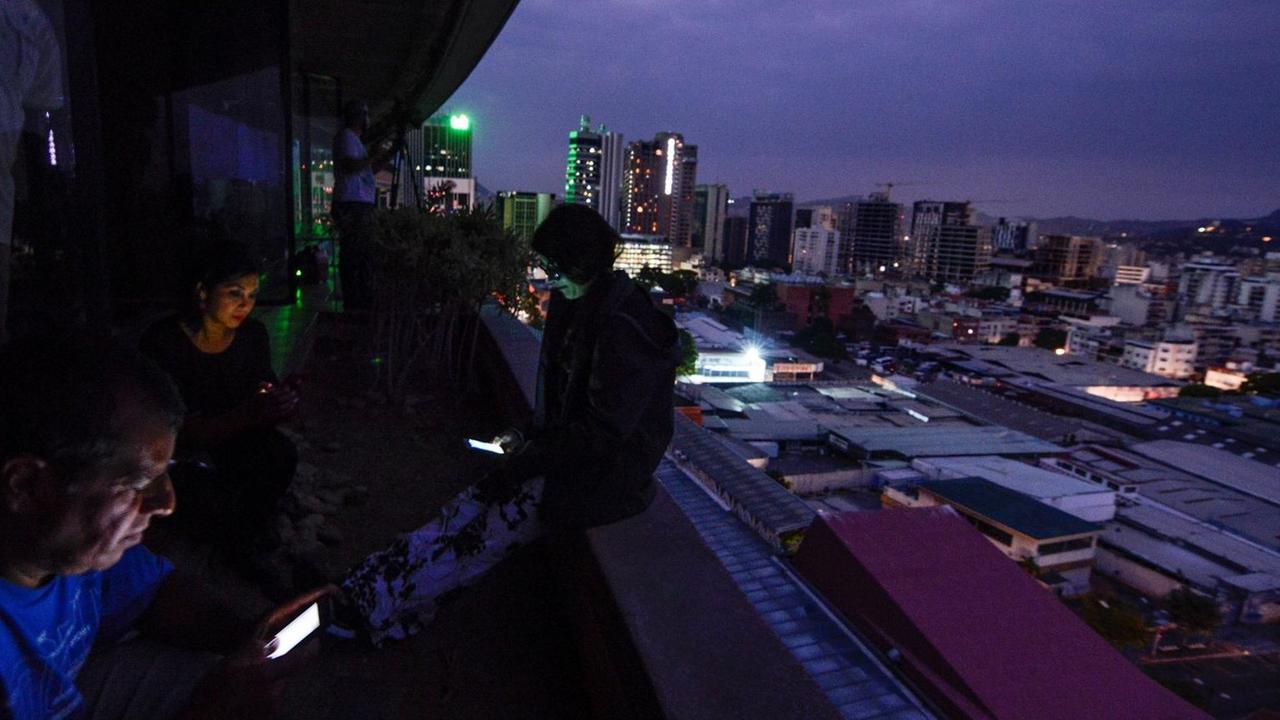 Menschen stehen mit beleuchteten Smartphones auf einer Terasse und blicken auf die Stadt Caracas. 
