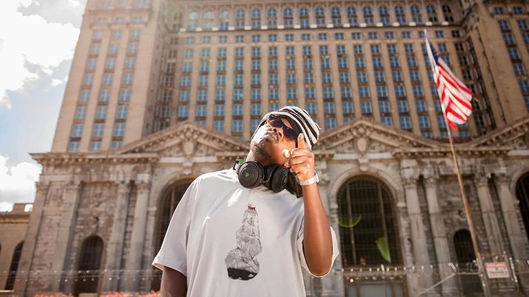 Ein Rapper mit ausgestrecktem linken Zeigefinger vor dem verwaisten Bahnhof Detroit, rechts weht die US-Flagge Ein Rapper mit ausgestrecktem linken Zeigefinger vor dem verwaisten Bahnhof Detroit, rechts weht die US-Flagge