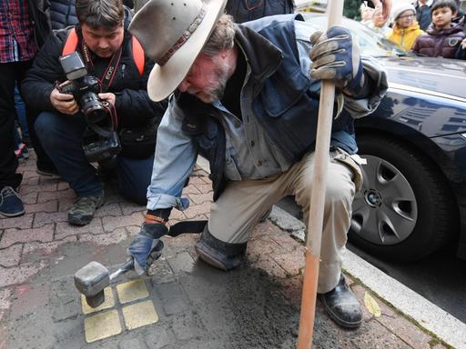 Gunter Demnig kniet auf dem Bürgersteig und klopft mit einem Hammer einen Stolperstein mit Messingplakette fest.