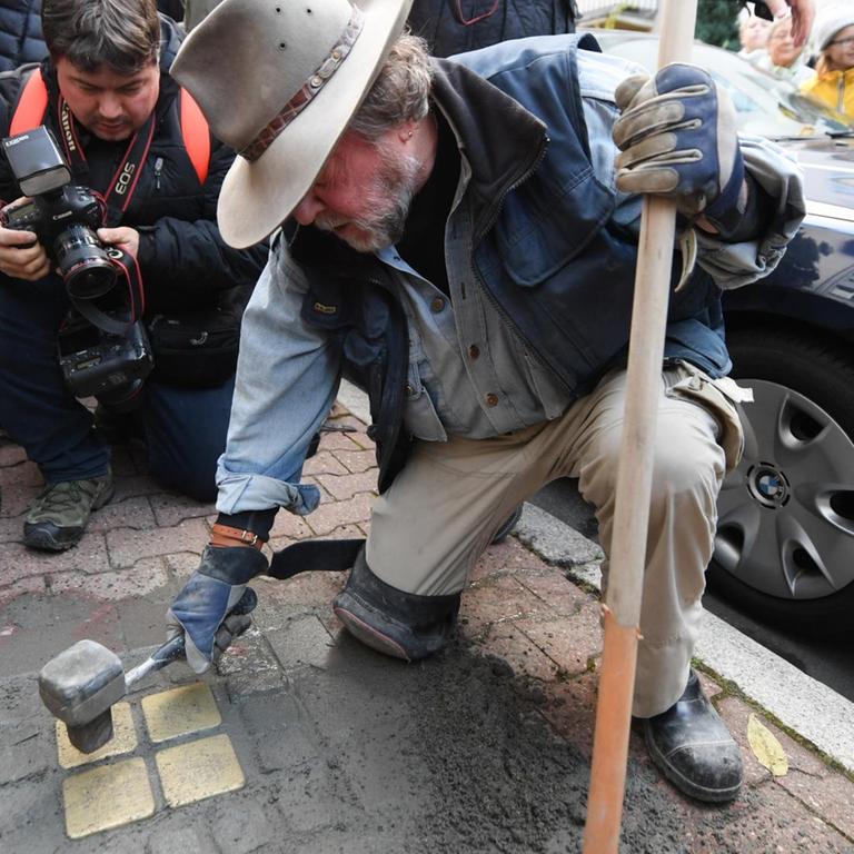 Gunter Demnig kniet auf dem Bürgersteig und klopft mit einem Hammer einen Stolperstein mit Messingplakette fest.