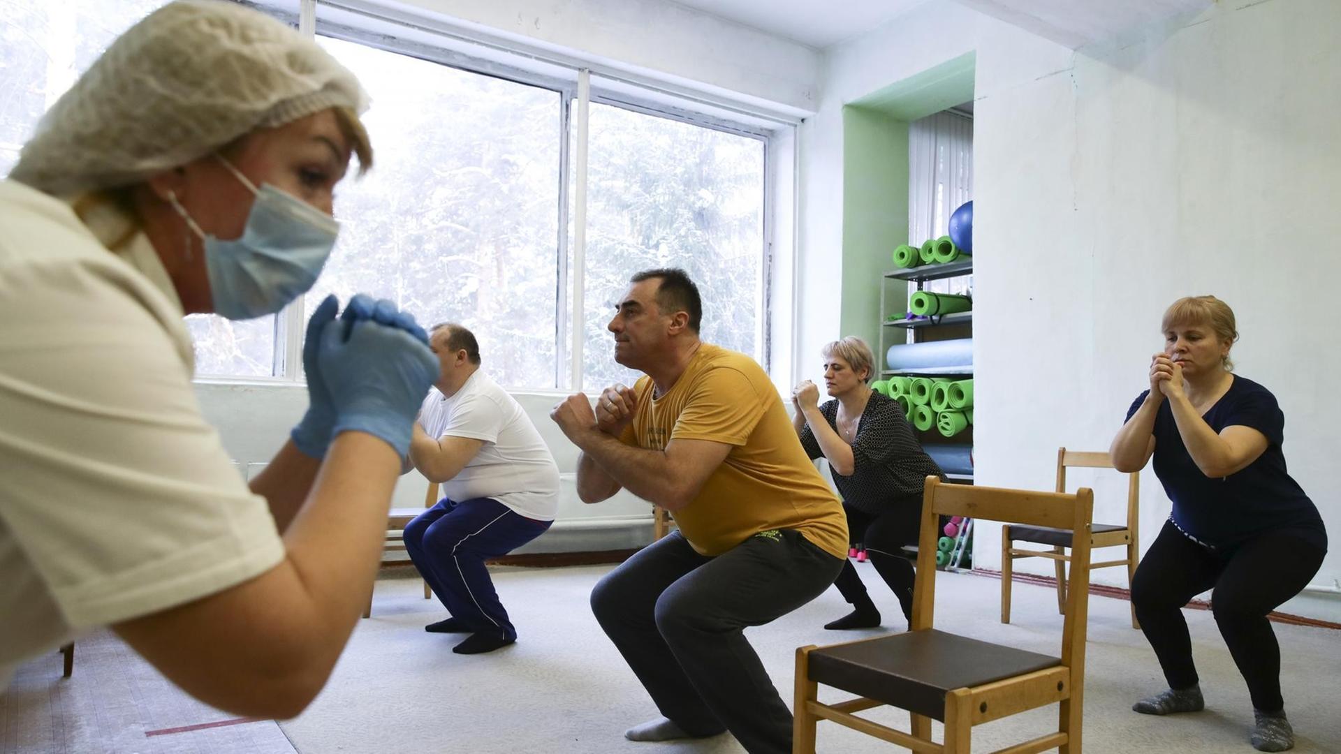 In einem Raum machen mehrere Menschen eine Kniebeuge. Vor ihnen steht eine Krankenschwester mit einem Mundnasenschutz. In einem Raum machen mehrere Menschen eine Kniebeuge. Vor ihnen steht eine Krankenschwester mit einem Mundnasenschutz.