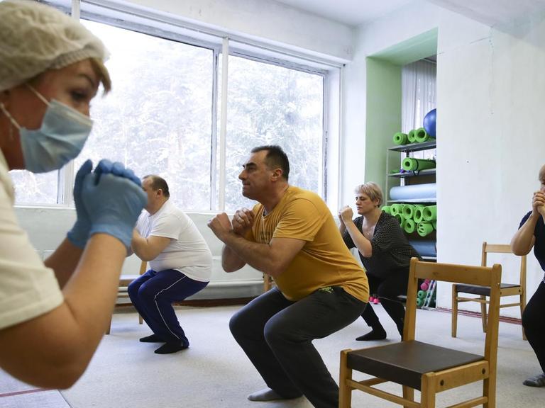 In einem Raum machen mehrere Menschen eine Kniebeuge. Vor ihnen steht eine Krankenschwester mit einem Mundnasenschutz.