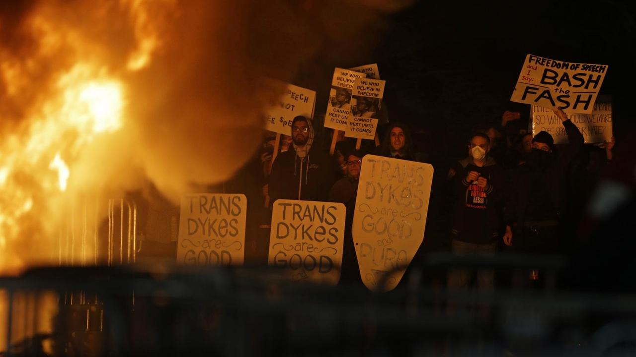 In der Dunkelheit sind Flammen zu sehen. Daneben stehen Menschen mit Protestplakaten. In der Dunkelheit sind Flammen zu sehen. Daneben stehen Menschen mit Protestplakaten.
