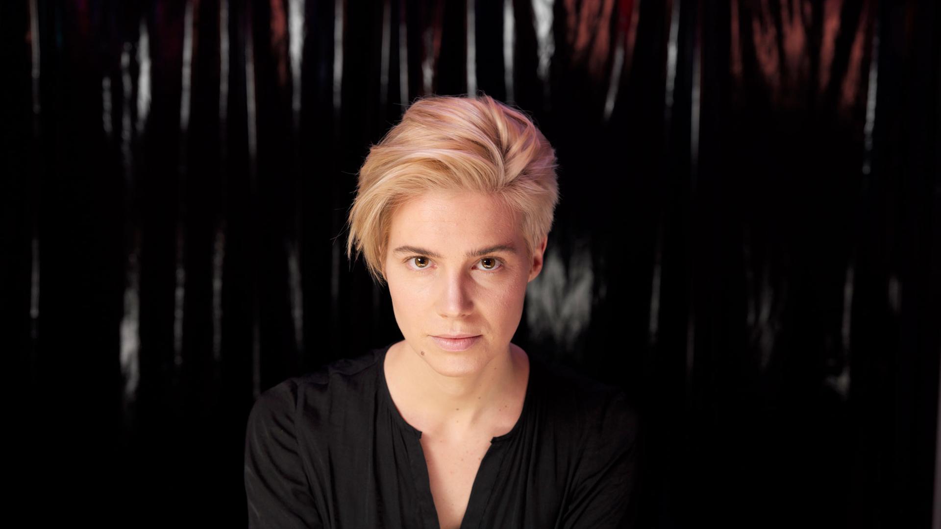 Eine junge Frau mit blonden kurzen Haaren trägt ein schwarzes Shirt und blickt in die Kamera. Der Hintergrund ist dunkel. Eine junge Frau mit blonden kurzen Haaren trägt ein schwarzes Shirt und blickt in die Kamera. Der Hintergrund ist dunkel.