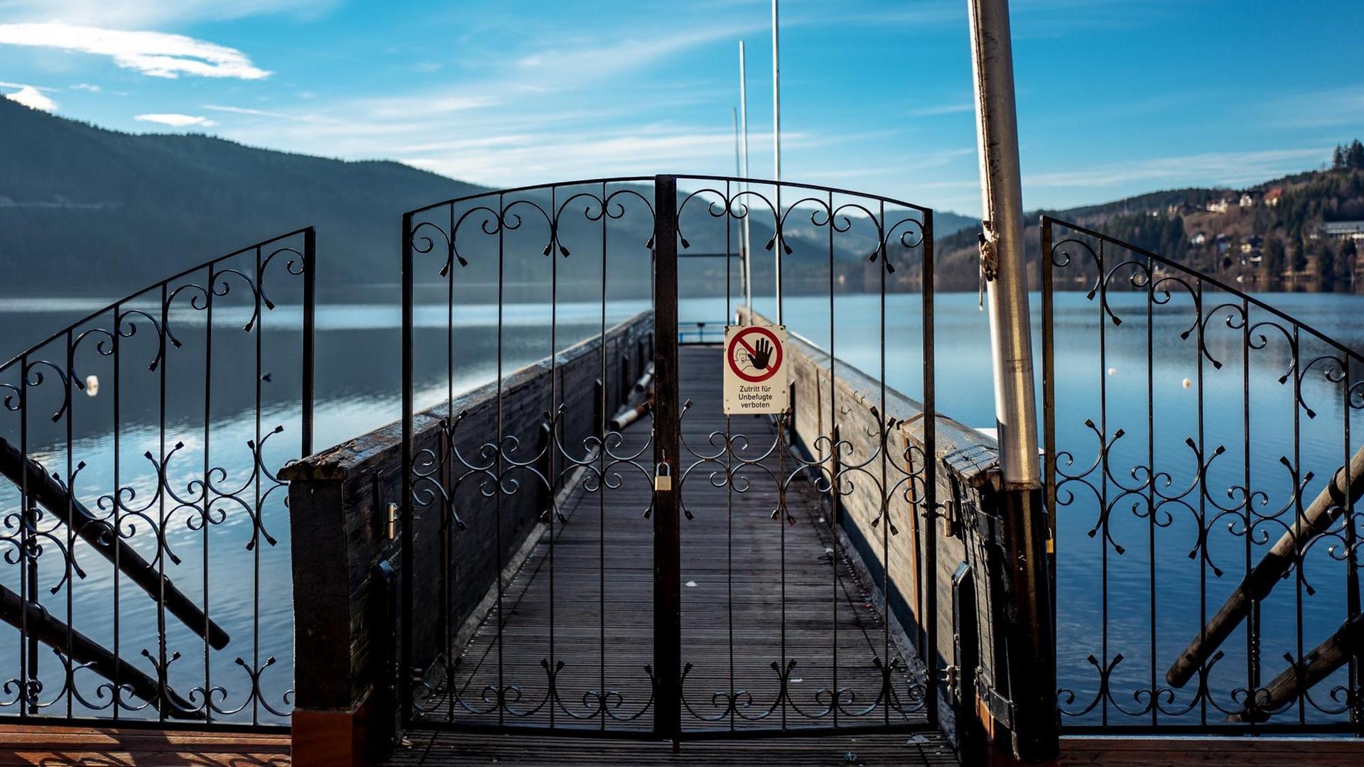 Blick auf einen Steg, der auf einen sonnenbeschienenen See führt. Davor ein verschlossenes schmiedeeisernes Tor mit einem Schild, auf dem steht "Zutritt für Unbefugte verboten". Blick auf einen Steg, der auf einen sonnenbeschienenen See führt. Davor ein verschlossenes schmiedeeisernes Tor mit einem Schild, auf dem steht "Zutritt für Unbefugte verboten".