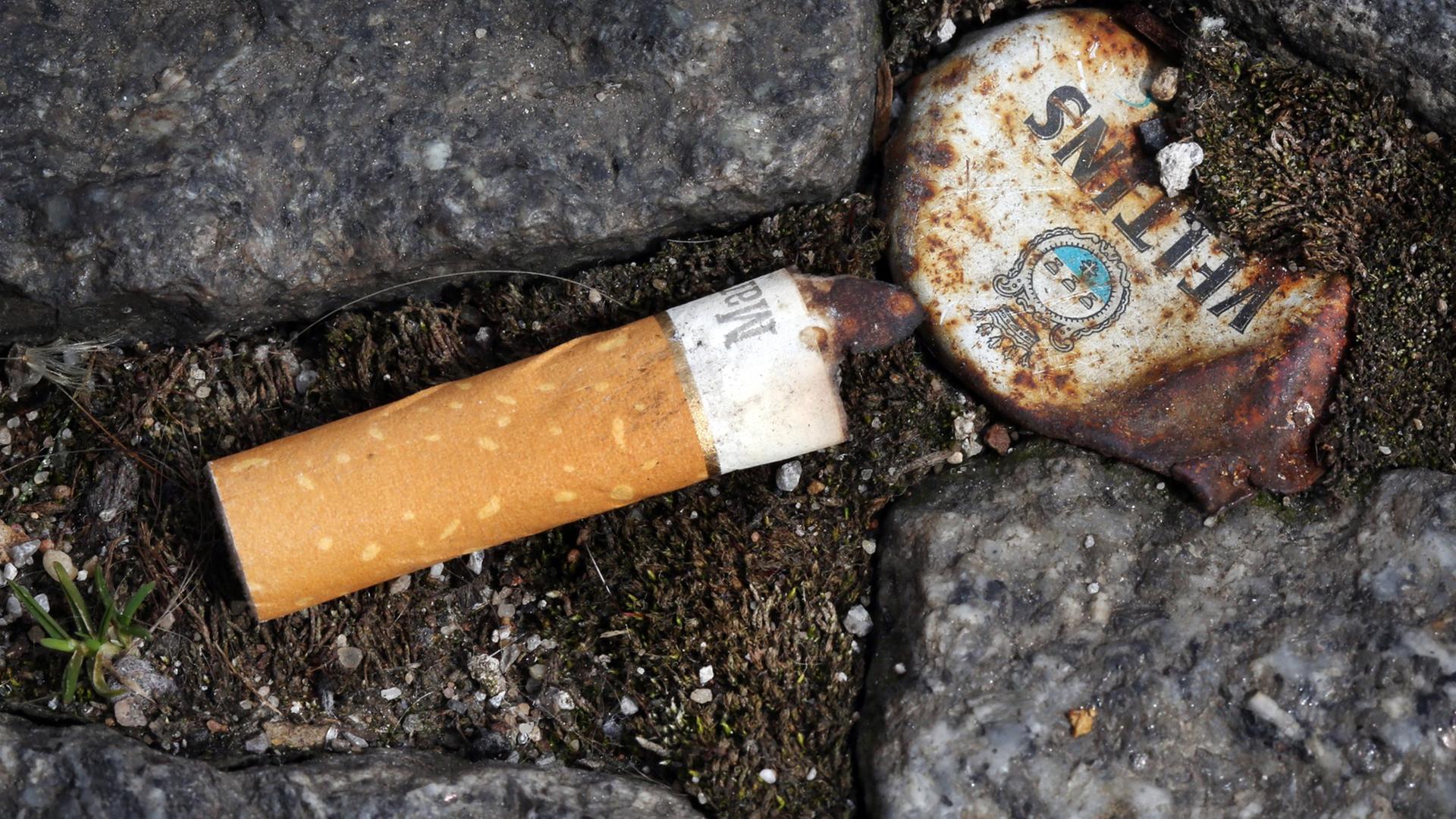 Ein Zigarettenstummel liegt neben einem rostigen Kronkorken zwischen Pflastersteinen auf dem Boden. Ein Zigarettenstummel liegt neben einem rostigen Kronkorken zwischen Pflastersteinen auf dem Boden.