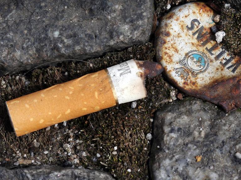 Ein Zigarettenstummel liegt neben einem rostigen Kronkorken zwischen Pflastersteinen auf dem Boden.