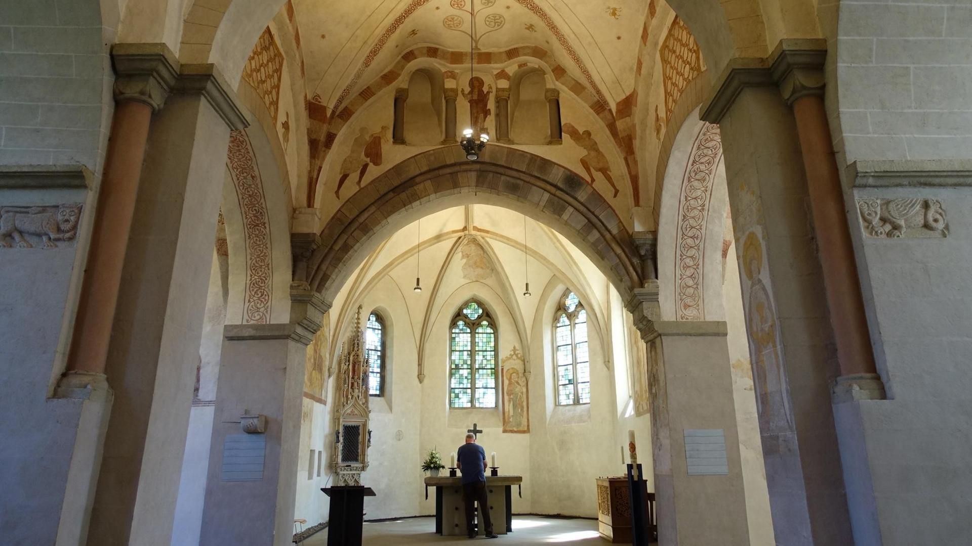 Der Innenraum der Stiepeler Dorfkirche, Blick zum Altar, vor dem ein Mann in dunkler Kleidung steht, mit dem Rücken zum Betrachter. An den sonst schlichten Stein-Wänden erkennt man restaurierte mittelalterliche Malereien in rostroter Farbe.