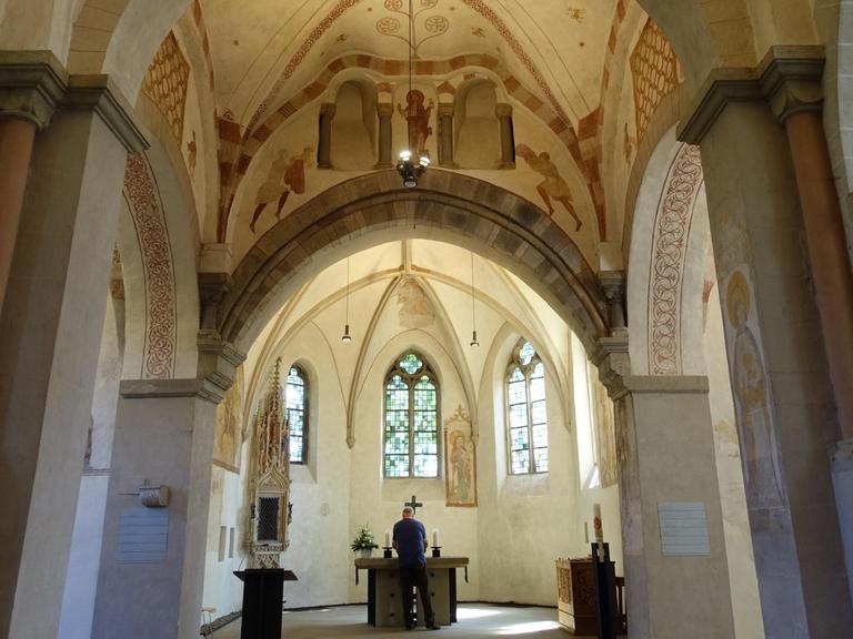 Der Innenraum der Stiepeler Dorfkirche, Blick zum Altar, vor dem ein Mann in dunkler Kleidung steht, mit dem Rücken zum Betrachter. An den sonst schlichten Stein-Wänden erkennt man restaurierte mittelalterliche Malereien in rostroter Farbe.