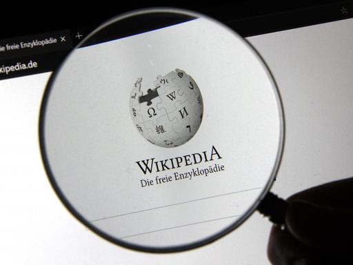 Durch eine Lupe ist das Logo der Online-Enzyklopädie Wikipedia zu sehen. 