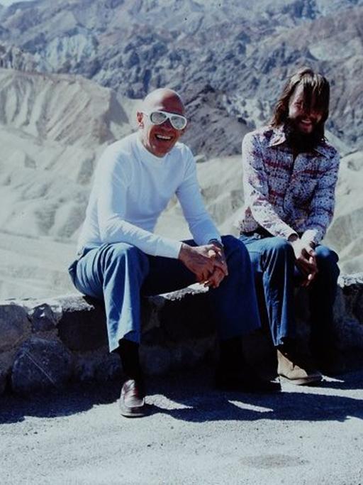 Am Rande einer Strasse auf einem kleinen Mäuerchen sitzen Foucault und Stoneman. Im Hintergrund die Berge vom Death Valley.