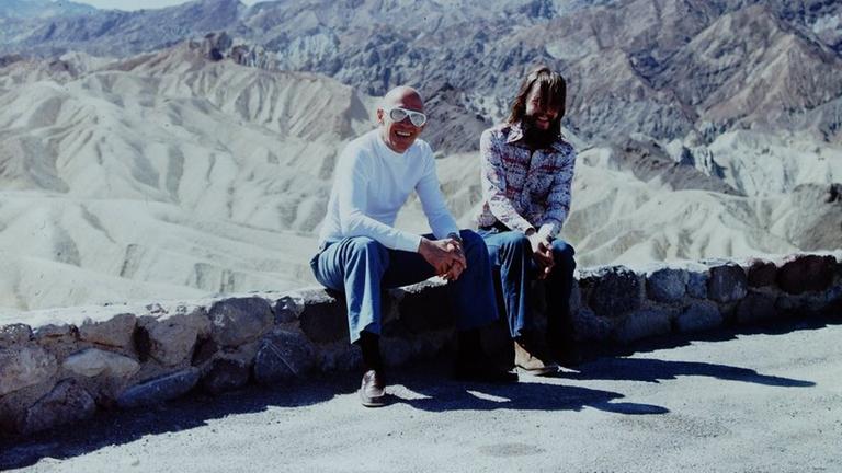 Am Rande einer Strasse auf einem kleinen Mäuerchen sitzen Foucault und Stoneman. Im Hintergrund die Berge vom Death Valley. Am Rande einer Strasse auf einem kleinen Mäuerchen sitzen Foucault und Stoneman. Im Hintergrund die Berge vom Death Valley.