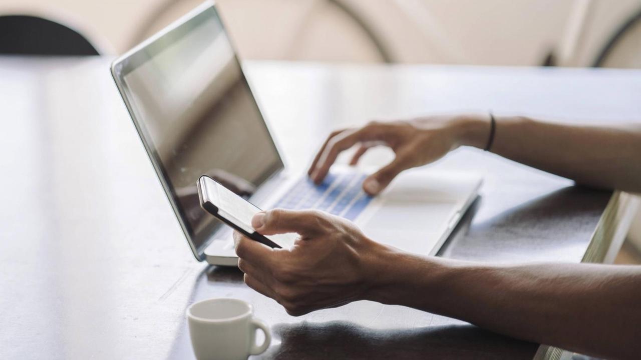 Jemand sitzt an einem Tisch vor einem Laptop und hält mit einer Hand ein Smartphone, im Hintergrund ist ein Fahrrad zu sehen, im Vordergrund eine Tasse. Jemand sitzt an einem Tisch vor einem Laptop und hält mit einer Hand ein Smartphone, im Hintergrund ist ein Fahrrad zu sehen, im Vordergrund eine Tasse.