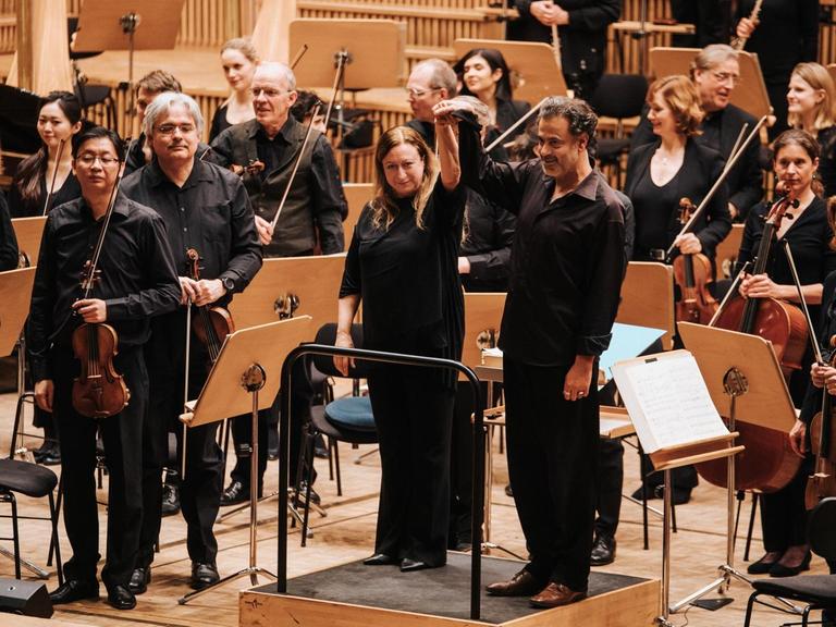 Der Komponist steht neben der Dirigentin vor dem Orchester