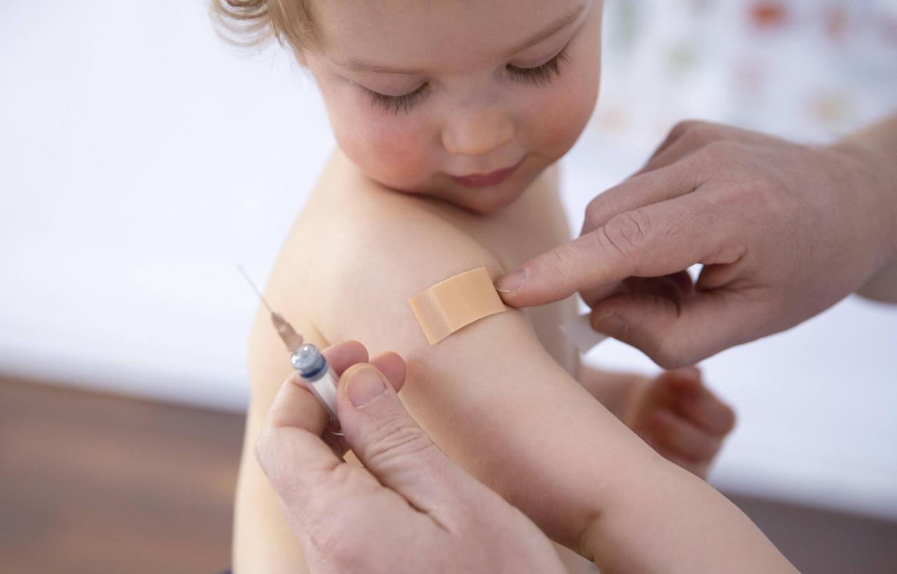 Impfung von einem Kind. Es schaut nach unten und die Arzthand klebt gerade ein Pflaster auf den Oberarm.