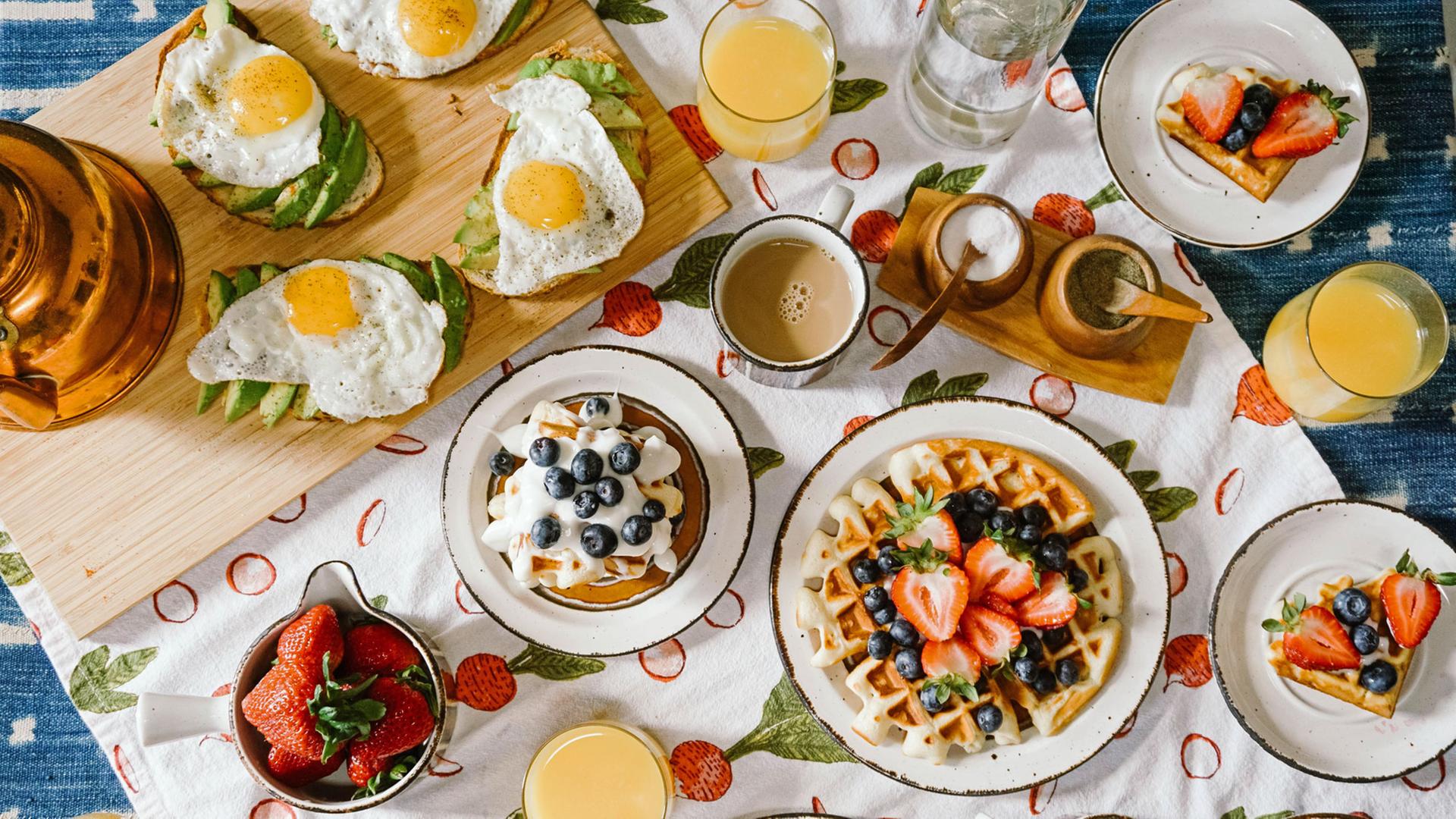 Blick von oben auf einen Tisch mit einer gefüllten Kaffeetasse, mit Spiegelei und Avocado belegten Broten und Waffeln mit Obst und Sahne. Blick von oben auf einen Tisch mit einer gefüllten Kaffeetasse, mit Spiegelei und Avocado belegten Broten und Waffeln mit Obst und Sahne.