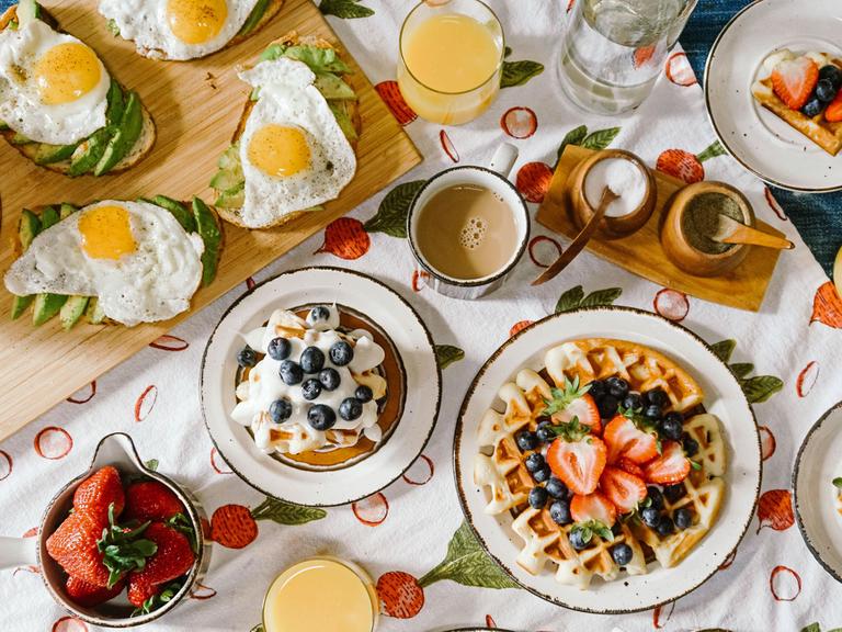 Blick von oben auf einen Tisch mit einer gefüllten Kaffeetasse, mit Spiegelei und Avocado belegten Broten und Waffeln mit Obst und Sahne.