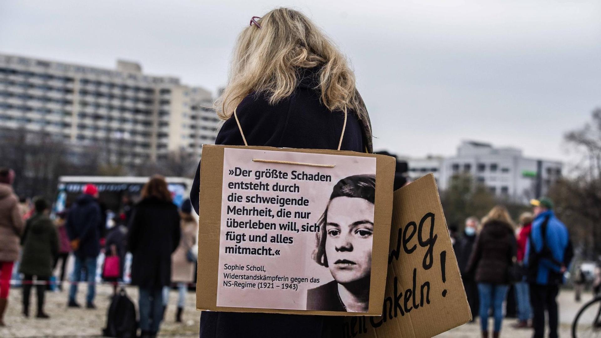 Rückansicht einer Frau auf einer Demo. Sie trägt um den Hals ein Schild mit dem Foto von Sophie Scholl, auf dem das Scholl zugeschrieben Zitat steht: "Der größte Schaden entsteht durch die schweigende Mehrheit, die nur überleben will, sich fügt und alles mitmacht." Rückansicht einer Frau auf einer Demo. Sie trägt um den Hals ein Schild mit dem Foto von Sophie Scholl, auf dem das Scholl zugeschrieben Zitat steht: "Der größte Schaden entsteht durch die schweigende Mehrheit, die nur überleben will, sich fügt und alles mitmacht."