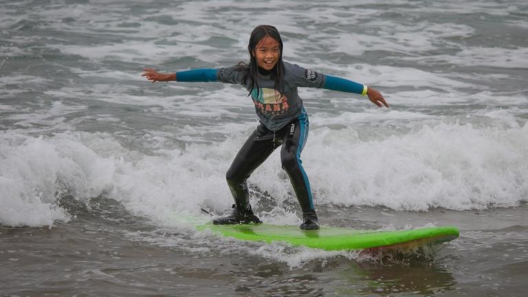 Ein Mädchen bei ihren ersten Surfversuchen, sie hält sich mit ausgebreiteten Armen auf dem Brett.