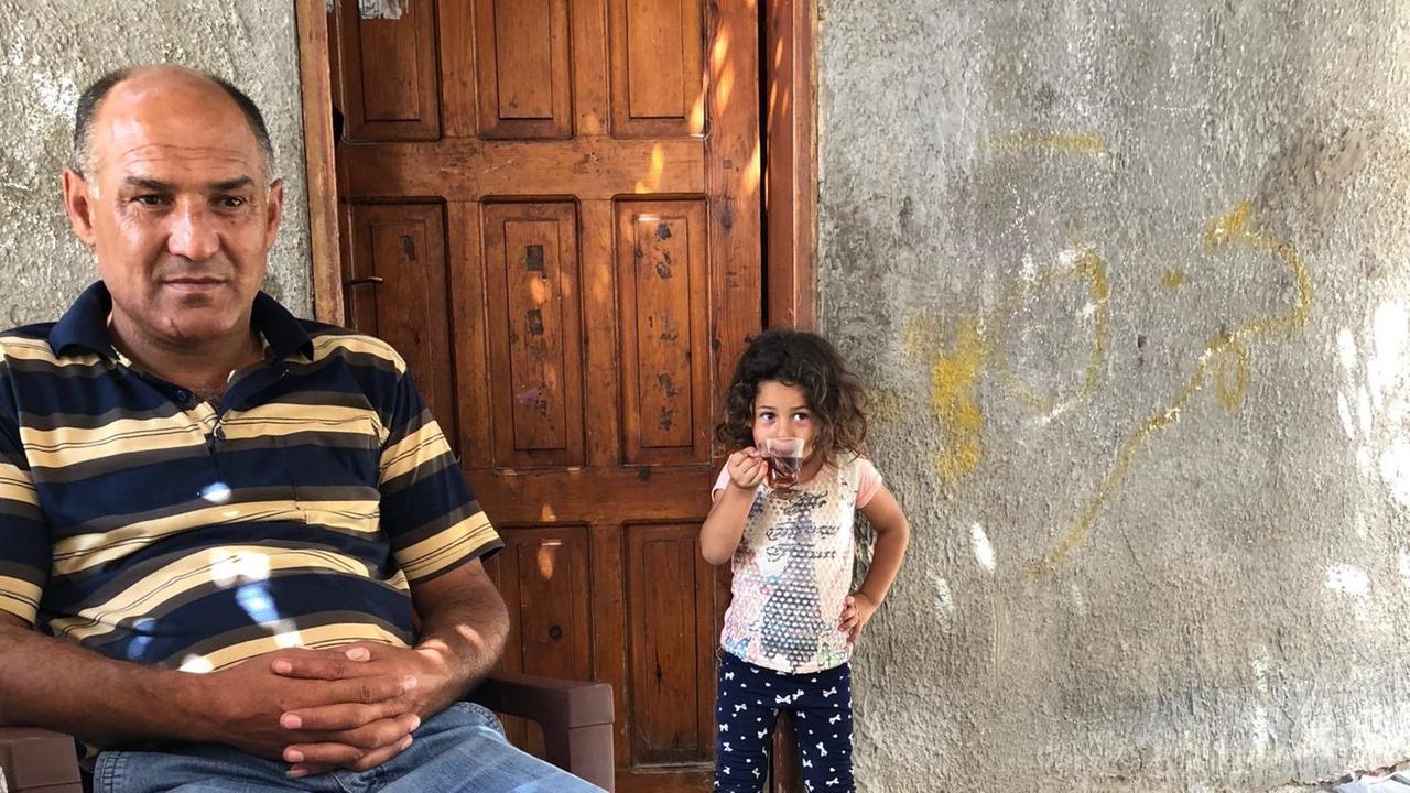 Raed Atamneh, Fahrer für Gäste im Gazastreifen, sitzt vor einer Tür im Hintergrund ein kleines Mädchen, das in die Kamera schaut