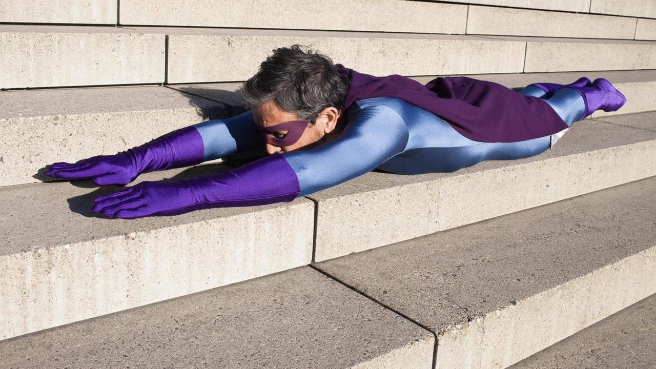 Ein Mann als Superheld verkleidet, mit violetter Augenbinde und Handschuhen, liegt auf einer Treppe auf dem Bauch, seine Arme nach vorn ausgestreckt. Ein Mann als Superheld verkleidet, mit violetter Augenbinde und Handschuhen, liegt auf einer Treppe auf dem Bauch, seine Arme nach vorn ausgestreckt.