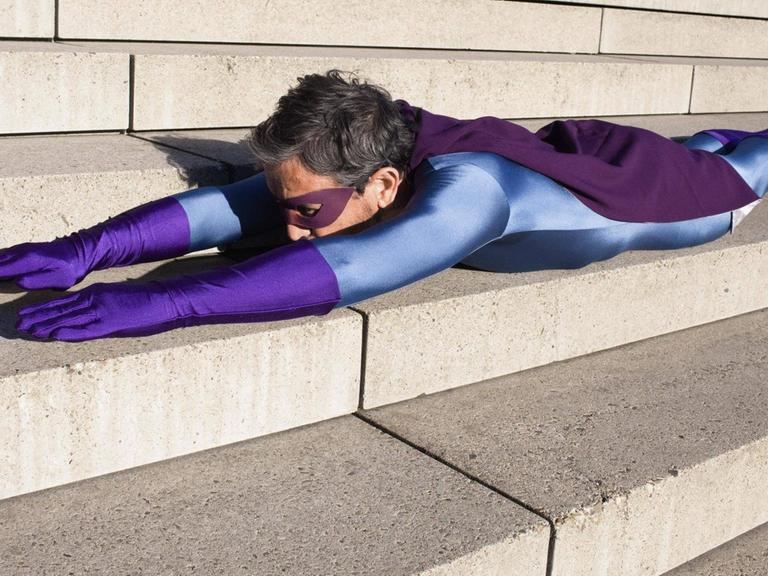Ein Mann als Superheld verkleidet, mit violetter Augenbinde und Handschuhen, liegt auf einer Treppe auf dem Bauch, seine Arme nach vorn ausgestreckt.
