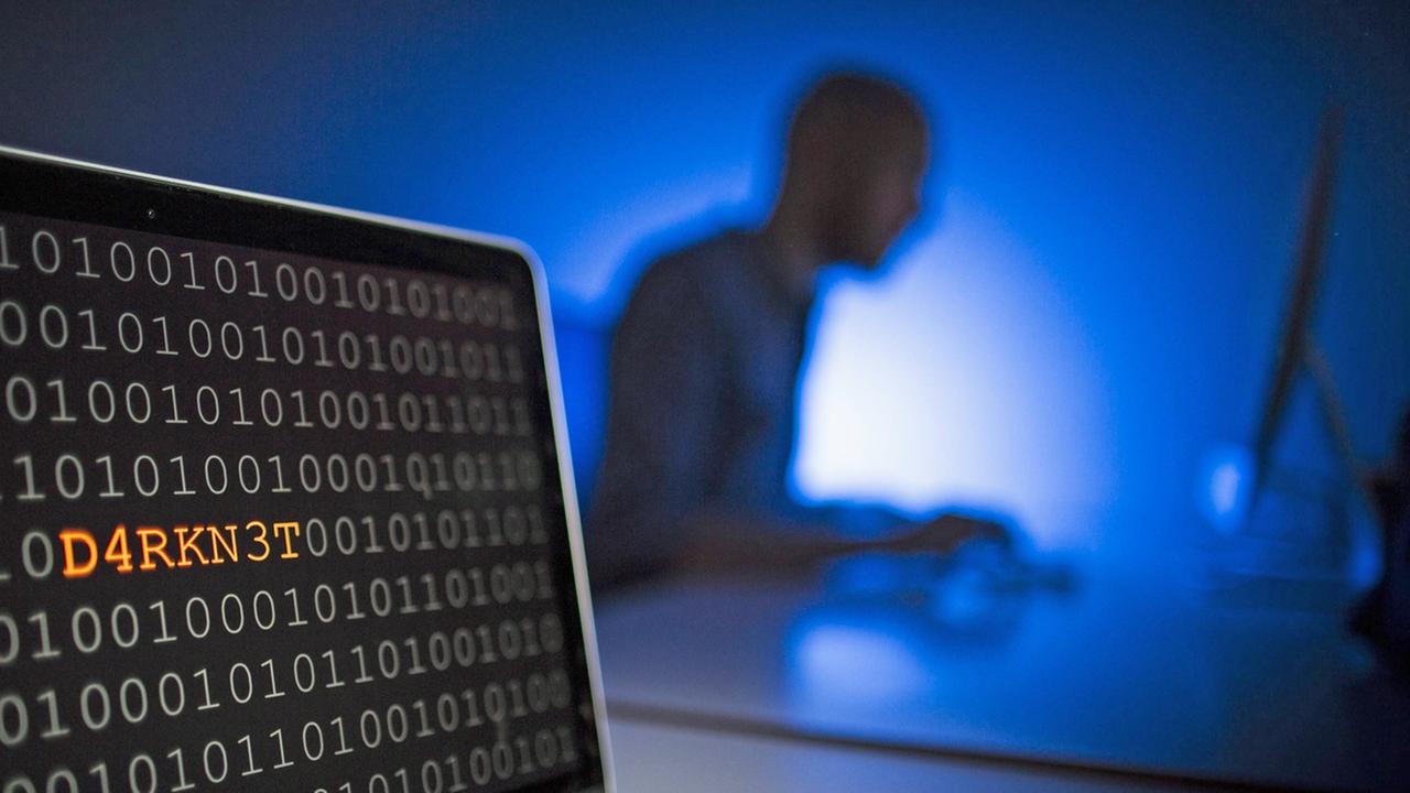 Nahaufnahme eines Laptops mit Binärcode, in dem das Wort "DARKN3T" zu lesen ist. Im Hintergrund sitzt ein Mann an einem Computer.