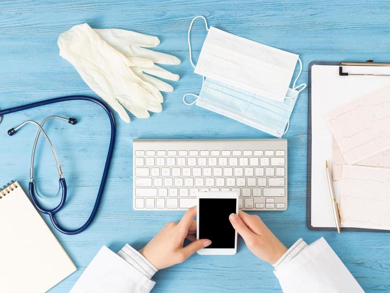 Der Schreibtisch einer Arztpraxis mit Handschuhe, Smartphone, Stereoskope, Maske und Papieren.