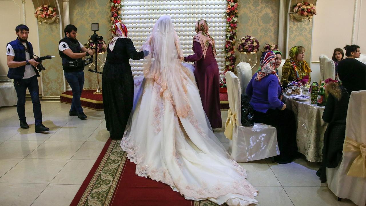 Eine Braut in weißem Kleid und mit langem Schleier wird von zwei Frauen zu ihrem Platz geführt. Links stehen Fotografen bereit, rechts sitzen Frauen an einem Tisch.