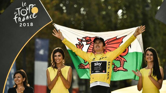 Tour-de-France-Sieger Geraint Thomas hält bei der Siegerehrung eine Flagge von Wales in den Händen, neben ihm stehen zwei klatschende Frauen in gelbern Kleidern.