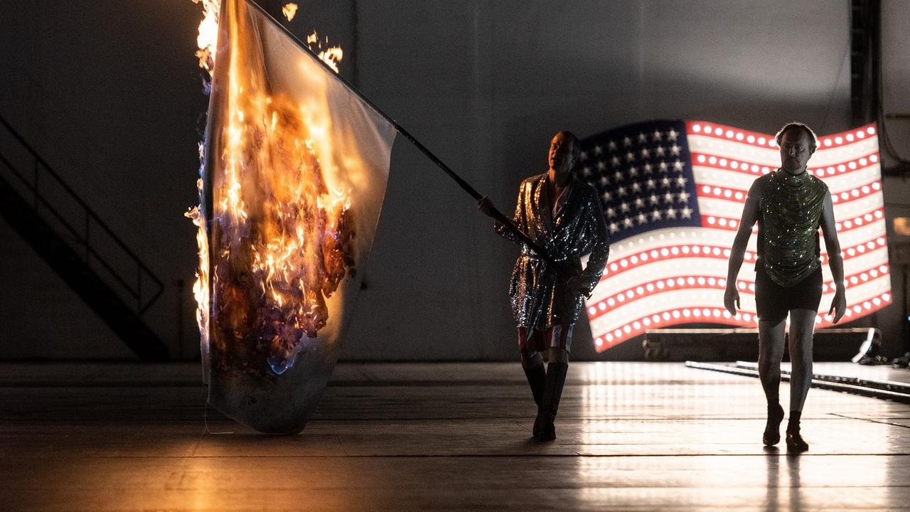 Die Schauspieler Matthias Klink und Georg Nigl in Frank Castorfs Inszenierung "molto agitato" an der Staatsoper Hamburg: Klink trägt eine brennende Fahne, im Hintergrund die US-amerikanische Flagge.