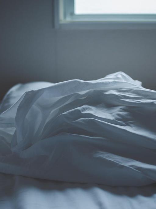 Ein unordentliches Bett mit weißen Laken. Durch das Fenster im Hintergrund fällt Licht aufs Bett.