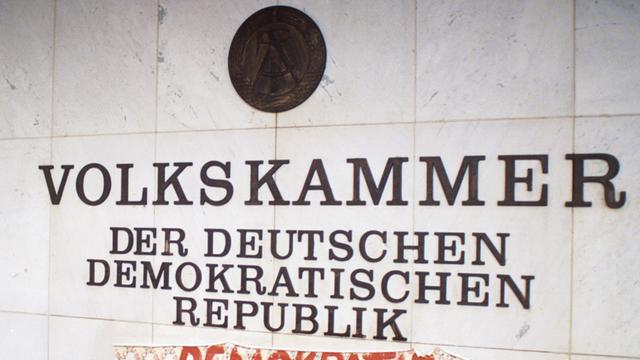 Ein Stück Tapete mit dem Spruch "Demokratie hier und jetzt" klebt am 04.11.1989 unter dem Schriftzug Volkskammer der Deutschen Demokratischen Republik am Palast der Republik in Ost-Berlin.