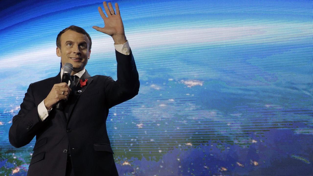 Macron steht lächelnd vor einer großen Videoleinwand mit der Erdkugel, lächelt, spricht und gestikuliert mit der Hand.