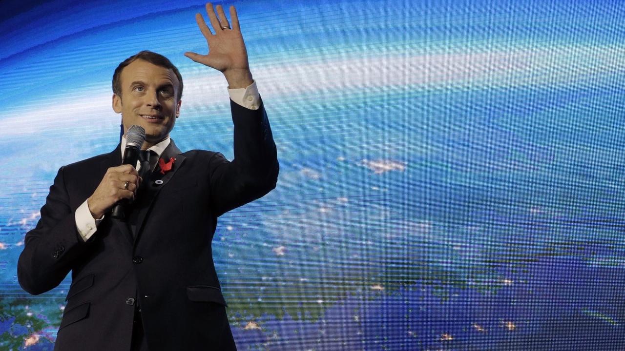 Macron steht lächelnd vor einer großen Videoleinwand mit der Erdkugel, lächelt, spricht und gestikuliert mit der Hand. Macron steht lächelnd vor einer großen Videoleinwand mit der Erdkugel, lächelt, spricht und gestikuliert mit der Hand.