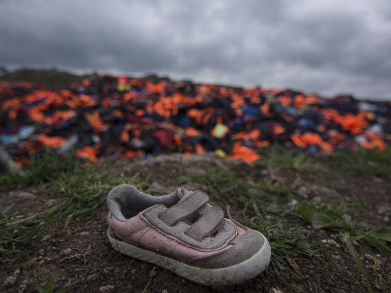 Tausende von Schwimmwesten, die von Flüchtlingen genutzt wurden, stapeln sich auf einer Wiese auf Lesbos. Im Vordergrund ein schmutzig-rosa Kinderschuh.