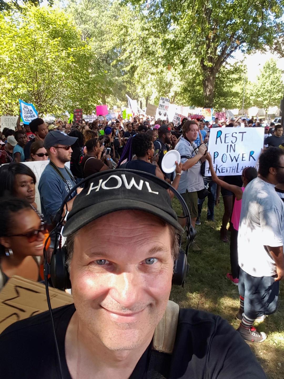 Der Autor Tom Schimmeck mit Käppi und Kopfhörern bei einer Demonstration in den USA lächelt in die Kamera.