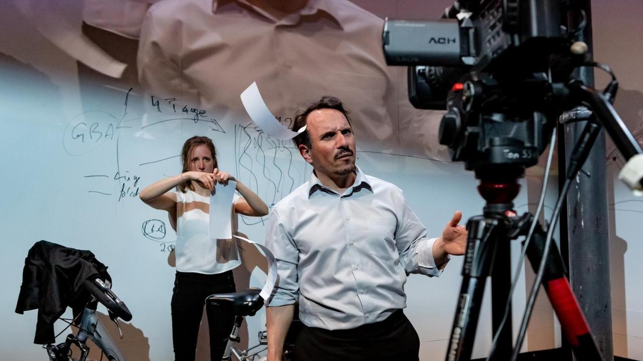 Foto von der Inszenierung: Eine Frau zerreißt Papier. Ein Mann steht vor ihr. Beide blicken in eine Kamera.