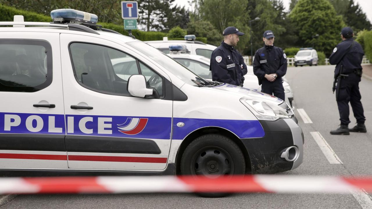 Das Bild zeigt ein Polizeiauto und drei Polizisten in schwarzen Uniformen hinter einem Absperrband auf einer Straße. Im Hintergrund sieht man Bäume und eine Hecke. Das Bild zeigt ein Polizeiauto und drei Polizisten in schwarzen Uniformen hinter einem Absperrband auf einer Straße. Im Hintergrund sieht man Bäume und eine Hecke.