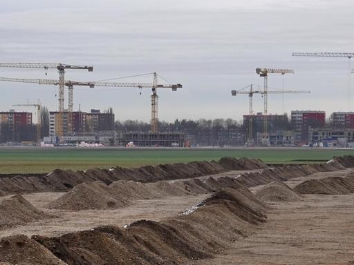Hinter einer grünen Wiese an einer Großbaustelle im Münchner Stadtteil Freiham stehen Kräne. Auf der neuen Bebauungsfläche ist der Bodenaushub in Reihen angeordnet.